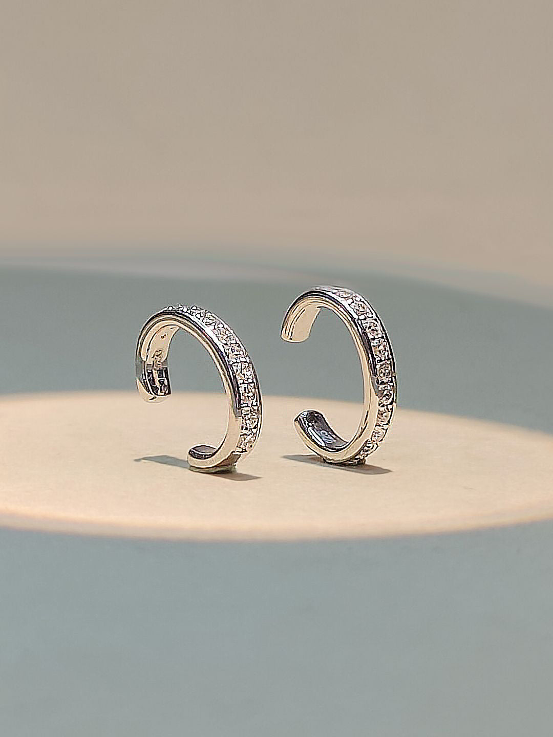 A pair of silver-toned open hoop earrings with rows of small clear stones, displayed on a light beige surface against a soft gray background.