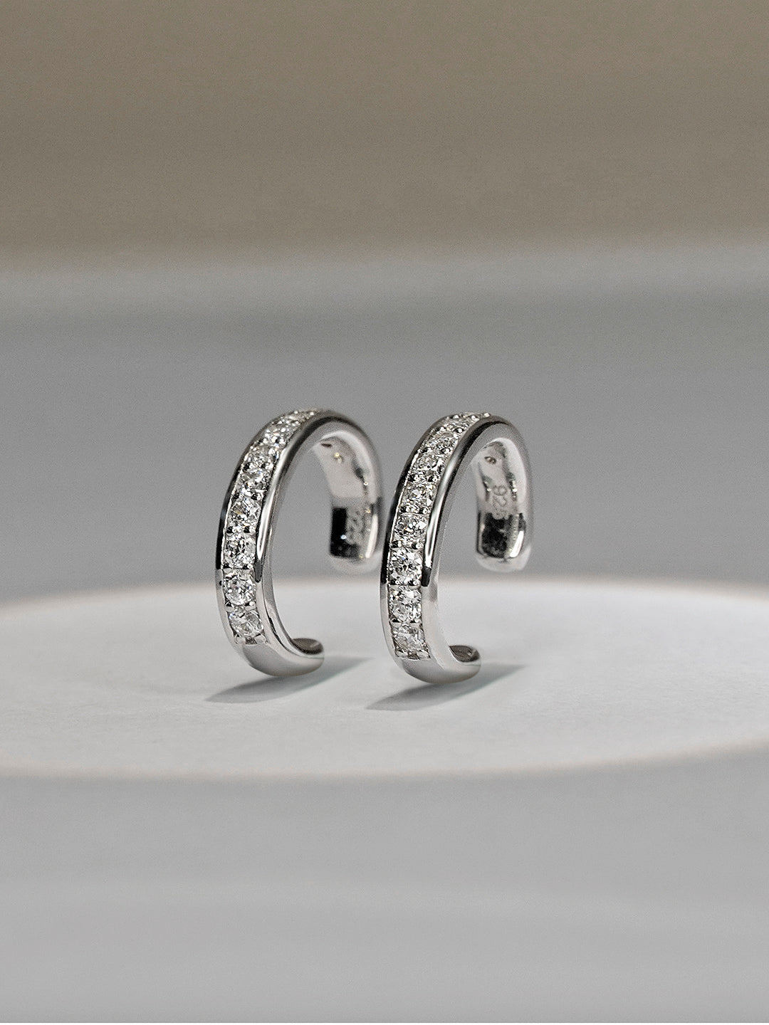 A pair of silver-toned curved hoop earrings with pavé-set clear stones, displayed on a light gray surface against a neutral gray background.