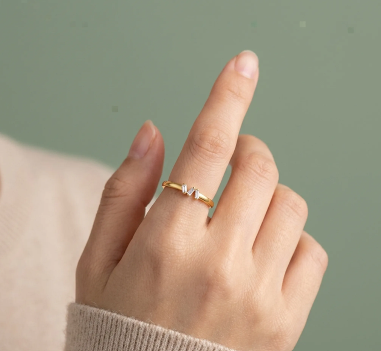 Delicate gold ring with a two-stone crystal accent, standing upright on a softly spotlighted neutral surface.